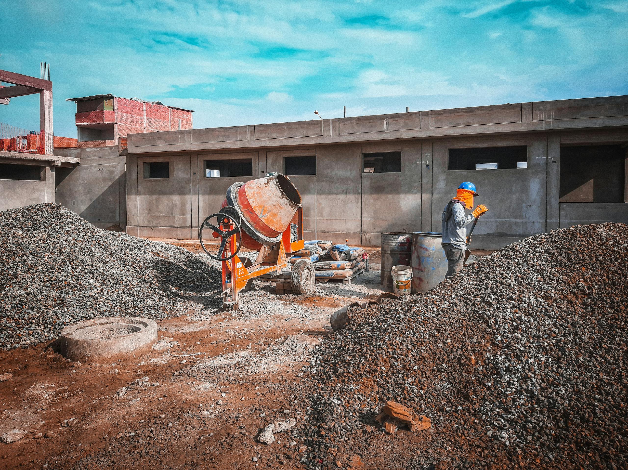 A construction site with a worker wearing safety gear next to a cement mixer and gravel piles.